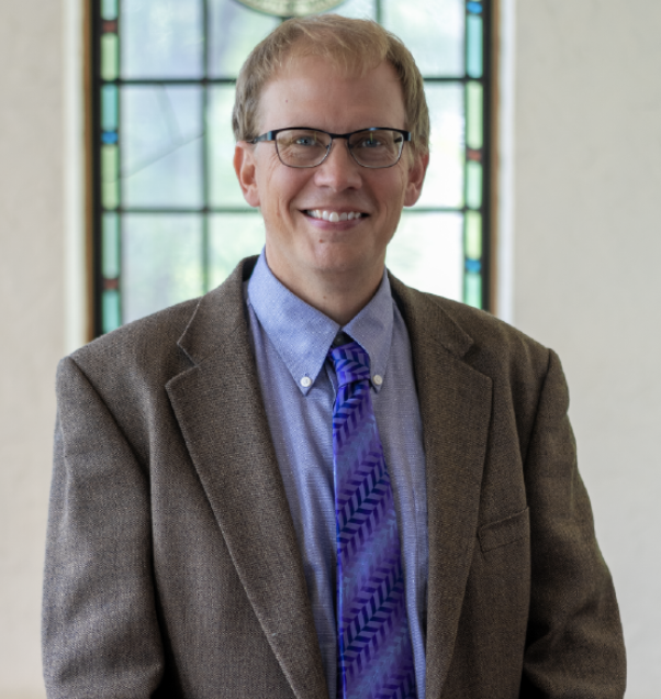 A man with short blond hair, glasses, and wearing a brown blazer, blue shirt, and purple tie is smiling. He stands indoors with a stained glass window in the background.